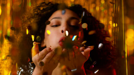Young-Woman-Celebrating-At-Party-Or-Club-Blowing-Handful-Of-Gold-Glitter-Towards-Camera